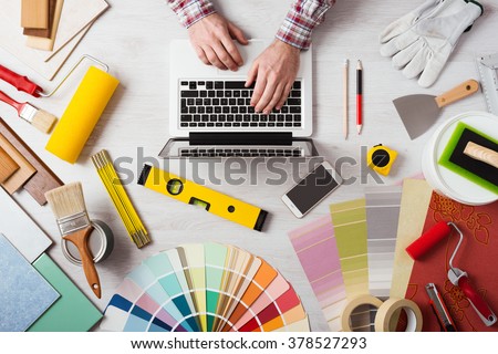 Professional decorator\'s hands working at his desk and typing on a laptop, color swatches, paint rollers and tools on work table, top view