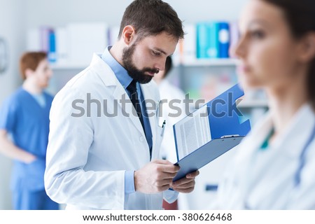 Confident doctor checking medical records on a clipboard in his office, medical staff on the background, healthcare concept