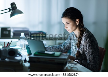 Pretty young student sitting at desk and doing her homework, she is connecting to the internet with a laptop