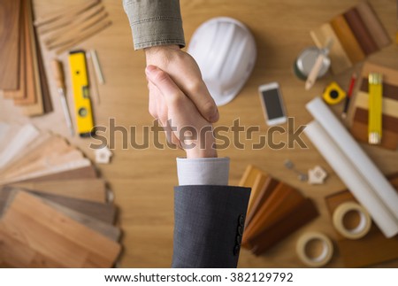 Construction engineer and businessman shaking hands top view close up, desktop with work tools and wood swatches on background