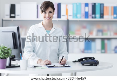 Smiling young female doctor working at the clinic reception, she is using a computer and writing medical reports