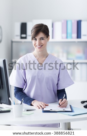 Smiling female doctor wearing a scrub and working at the hospital reception, she is writing a medical report on a clipboard and scheduling appointments
