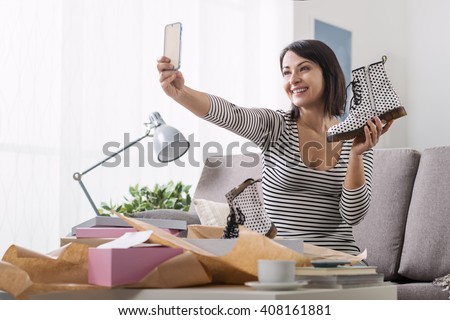 Smiling woman unboxing a postal parcel and taking a selfie with her new purchases using a smartphone