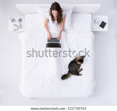 Young happy woman relaxing in her bed with her pet, she is connecting with a laptop, top view