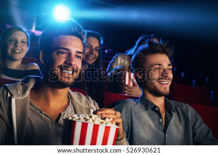 Young people sitting at the cinema, watching a movie and eating popcorn, two smiling men on foreground