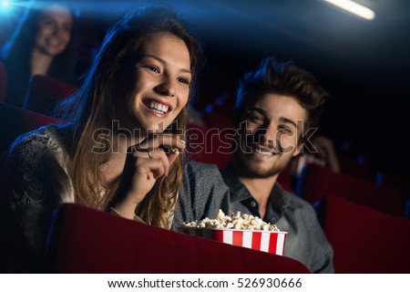 Young loving couple at the cinema watching a movie and smiling, people sitting on background