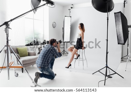 Professional photo shooting at the studio: a beautiful young model is smiling and posing; the photographer is taking pictures with a digital camera