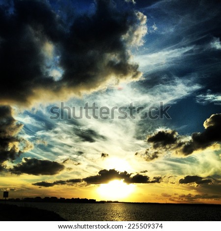 A cloudy sky with the sun setting over the silhouette of a city and water.