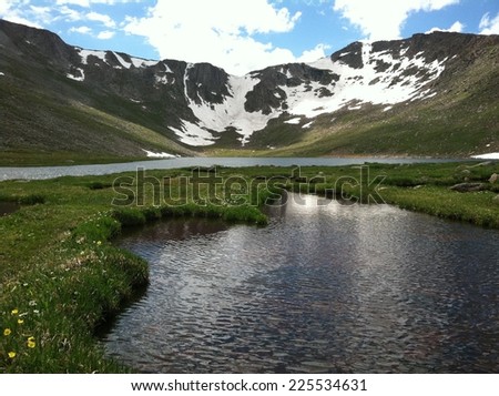 A body of calm water with green growth at the base of snow covered mountains.
