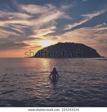 Woman wading out to sea at sunset towards a small island.