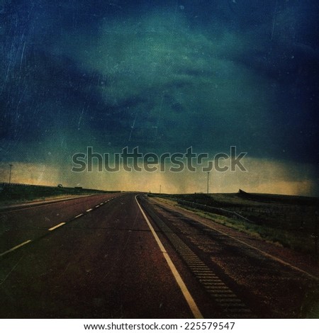 A deserted highway bordered by green fields leading to the edge of a cloudburst.