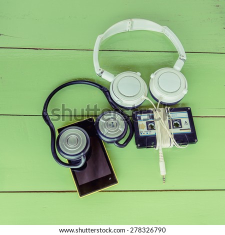 concept photo of smartphone with Bluetooth headset and  cassette tape with wire headphone. Smartphone cassette tape and headset put on green color wood board.