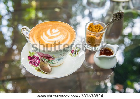 latte coffee with brown sugar and syrup jar on the glass table. The table surface has bokeh reflected light from tree shadow