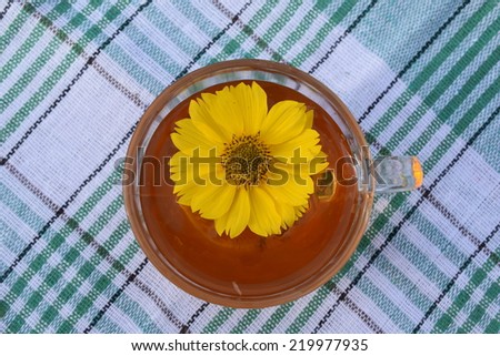Tea in a glass cup on a checkered white and green fabric and yellow flower in a bowl - top view