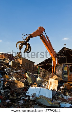 An orange hydraulic excavator with a scrap metal grab attached in the process of dropping metal onto the scrap heap set against a blue sky.