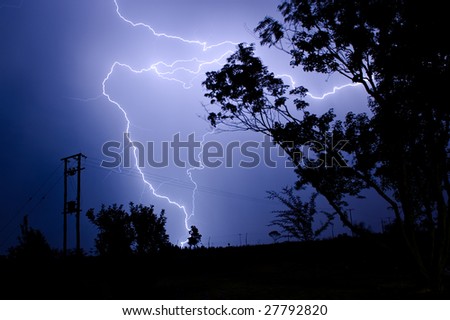 A massive branched lightning strike at night hitting the ground near high voltage power lines and silhouetted trees during a summer thunderstorm