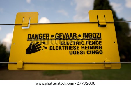 A bright yellow electric security fence warning sign posted in three languages, english, afrikaans and zulu, warning trespassers of possible high voltage electric shock.