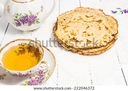 Hot tasty baked russian pancakes with tea in an antique thin porcelain painted cup and teapot on a white wooden background with lace cotton napkin. Holiday table for Shrovetide. Delicious breakfast