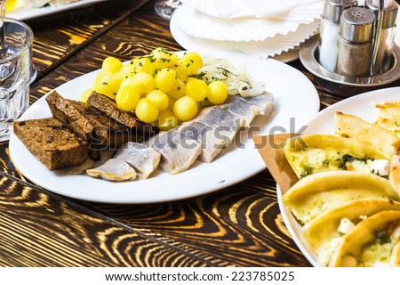 Dinner in russian restaurant - small fried potato dusted with greens and salted herring fish with bread toasted on a white plate on a brown wooden table