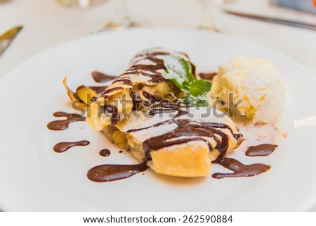 Appetizing apple strudel pie with ice-cream ball, mint leaf and chocolate drops on a white plate in a restaurant