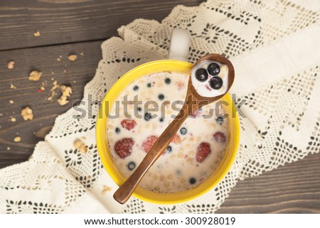 Muesli in a yellow ceramic cup with milk and berries on a dark wooden table with lace cotton napkin and wooden spoon