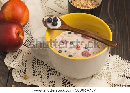 Muesli in a yellow ceramic cup with milk and berries on a dark wooden table with lace cotton napkin, peaches and wooden spoon