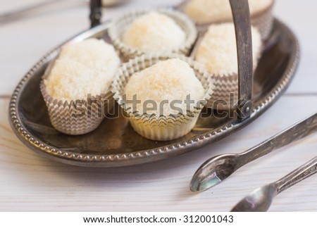 Coconut protein cupcake sweets in a paper pack on a silver plate with silver spoons and tongs on a grey napkin on a dark grey wooden background