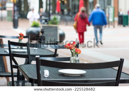 A couple walk hand in hand along an outdoor mall. In the foreground is a black metal table with a simple arrangement of roses with a white napkin and plates.