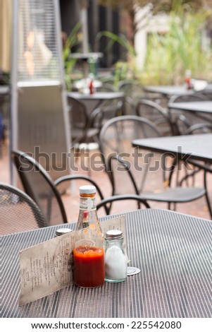 A metal outdoor restaurant table with a ketchup bottle, salt shaker, and menu. An outdoor heater and other tables are in the background.