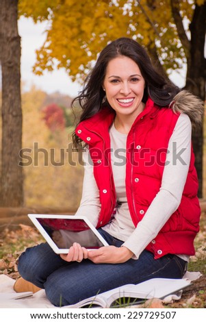 A pretty lady sits outside next to a tree and on a blanket. She has study materials including a book, notepad, and tablet. Fall colors. She is wearing a red vest, white shirt, and blue jeans.