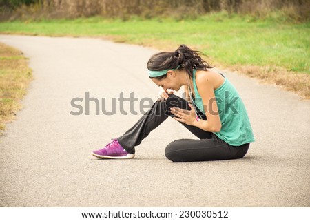 An athletic woman holds her knee in pain while sitting on a running path outside.