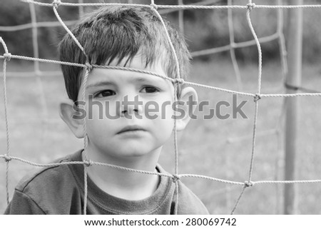 A black and white image of a little boy with his face in a net. He is making a serious and thoughtful face. Close up. Horizontal.