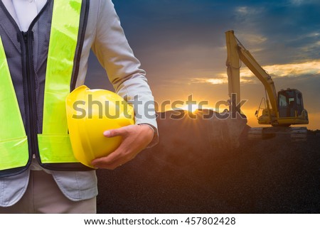 Engineer or Safety officer holding hard hat with excavator machine at construction site on sunset time is background.