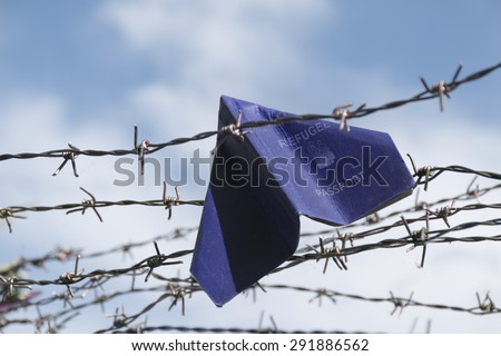 refugee labeled passport folded as a paper plane get stuck in the barbed wire of the border against the blue sky with white clouds, copy space