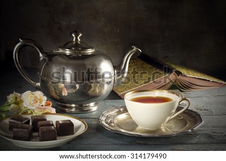 good old tea time, black tea in a porcelain cup, old-fashioned silver teapot, chocolate cookies and a good book on a rustic wooden table, copy space in the dark brown background