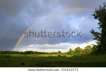 Double rainbow in the cloudy sky after a thunderstorm over a wide green country landscape with horse pasture, fields and trees, an idyllic rural background with copy space