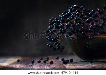 Black elderberries (Sambucus nigra) in a bowl and some berries on a rustic wooden table against a dark background with copy space, vintage still life, selected focus and very narrow depth of field