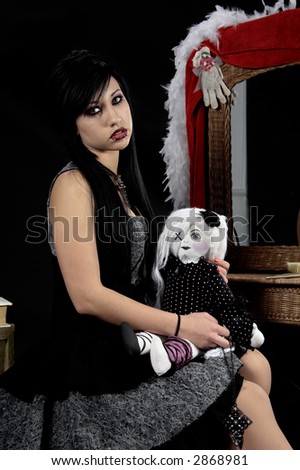 Beautiful raven haired Gothic Vampire Slayer in a black and grey dress and striking green eyes. sitting at her vanity with a doll in her arms and a cross necklace in her hand.