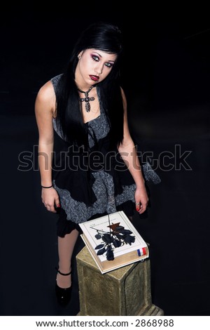 Beautiful raven haired Gothic Vampire girl in a black and grey dress and striking green eyes standing in front of a bible. Shot on black.
