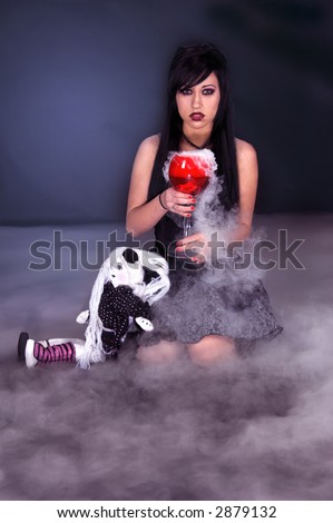Beautiful raven haired Gothic Vampire girl in a black and grey dress sitting in moonlight and fog with a goblet of witches brew and a doll.