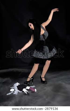 Beautiful raven haired Gothic Vampire girl in a black and grey dress dances over a doll laying on the fog