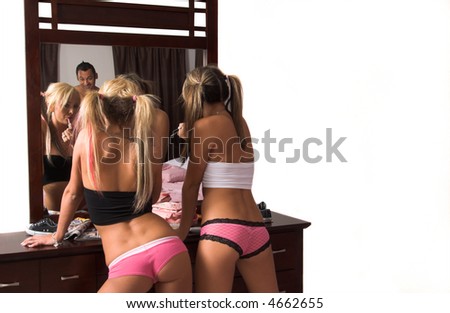 Two sexy young women applying lipstick in their bedroom with a man standing behind her watching her in the mirror