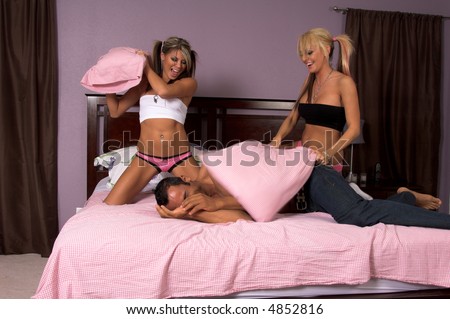 Two girls in lingerie having a pillow fight against a man on a bed