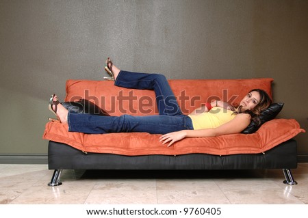 Pretty young Mexican woman in jeans and a yellow wife beater casually laying on a black and orange couch