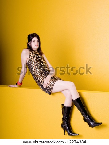A beautiful and hip young woman in a black and gold Couture dress sitting on a yellow wall