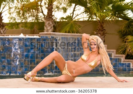 Sexy young bikini model with golden blonde hair wearing a gold swimsuit and high heel shoes in front of a pool fountain