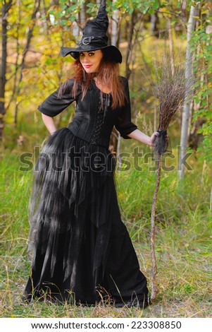 young long haired woman wearing black dress, black corset and witch hat, broom in hands, looking at camera