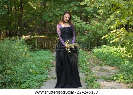 young woman in purple blouse, black floor-length dress and black corset holding wild flowers, looking downwards