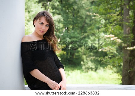 Semi-smiling long haired young woman in black loose knit dress leaning against a white pillar and looking at camera, trees in blurry background