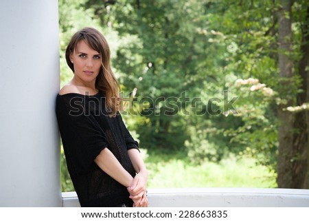 Long haired young woman in black loose knit dress leaning against a white pillar and looking at camera, trees in blurry background